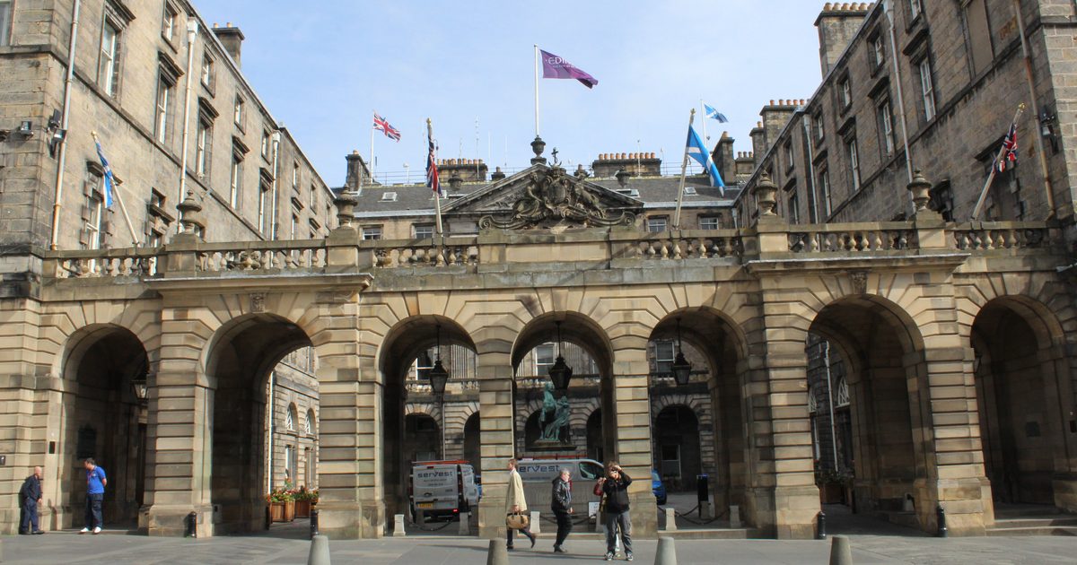 La fachada porticada del Edinburgh's City Chambers en el Royal Mile, siete arcos de piedra bajo una balaustrada y banderas, con el patio detrás